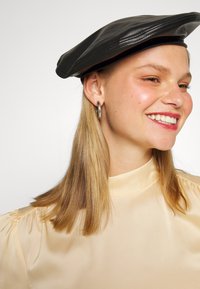 Boina de couro preto com topo plano e bordas costuradas, usada com uma blusa de seda bege. Modelo com cabelo loiro liso e brincos de argola metálicos.