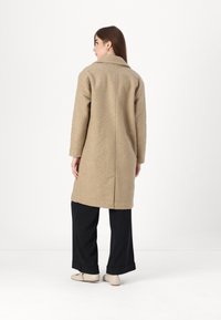 Beige ullblandingsfrakk med klaffekrage, lange ermer og rett passform, kombinert med svarte vide bukser og lyse sko.