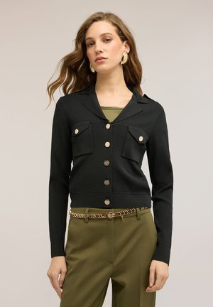 Donna con capelli lunghi e ondulati che indossa un cardigan nero con bottoni, una maglia verde oliva, pantaloni verde oliva e una cintura a fantasia, in piedi davanti a uno sfondo semplice.