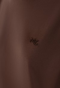Tissu marron au toucher lisse avec un logo en relief subtil "MG" près du coin inférieur droit. Gros plan mettant en avant les détails.