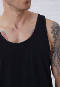 Zwarte katoenen tanktop met een ronde halslijn, met een gladde textuur en brede armsgaten. Decoratieve tatoeages gedeeltelijk zichtbaar op de huid.