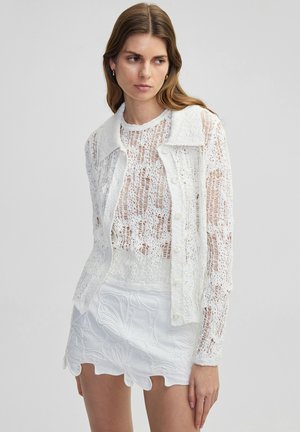 Cardigan bianco in crochet con colletto, chiusura con bottoni e texture trasparente, abbinato a una gonna mini bianca strutturata con ricami floreali.