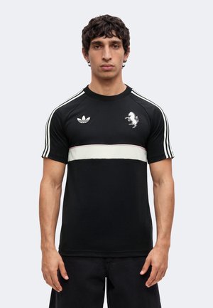 Man draagt een zwart Adidas T-shirt met een witte horizontale streep, drie witte schouderstrepen en twee logo’s op de borst, kijkt recht vooruit.