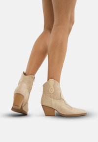 Botines de ante beige con puntera afilada, costuras decorativas y acentos de tachuelas; tacón bajo apilado; cierre de cremallera lateral.