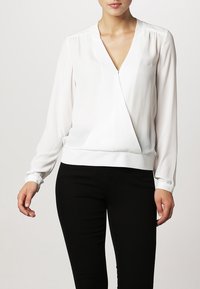 Weiße Wickelbluse aus leichtem Stoff, mit langen Ärmeln, tiefem V-Ausschnitt und schmalem Saum, kombiniert mit schwarzen Hosen.