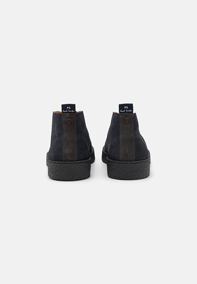 Zapatos de ante azul marino oscuro con una suela negra texturizada, que presentan un panel lateral liso, acentos en contraste y una pestaña con el logotipo en el talón.