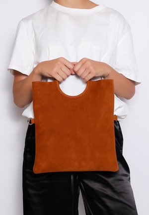 Personne en chemise blanche tenant un sac fourre-tout en suède couleur rouille avec des poignées circulaires devant un fond uni.