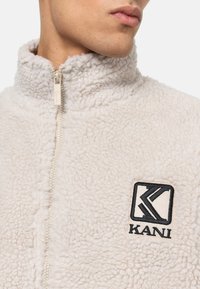 Ljusbeige fleecejacka med hög krage och halvhake. Har en svart broderad logotyp på vänster bröst. Mjukt, strukturerat tyg.