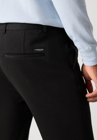 Pantalon noir avec une coupe ajustée, une texture lisse et une poche arrière avec une étiquette de marque. La taille est discrète, se fondant dans le tissu.