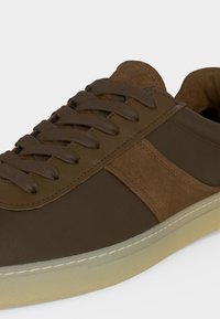 Καφέ αθλητικό παπούτσι με πάνελ από σουέντ και ύφασμα, καφέ κορδόνια που ταιριάζουν και ανοιχτό μπεζ σόλα, εμφανιζόμενο σε κοντινή γωνία.