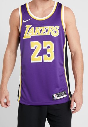 Muž nosící fialový basketbalový dres Los Angeles Lakers s číslem 23 a černé šortky, stojící na jednobarevném pozadí.