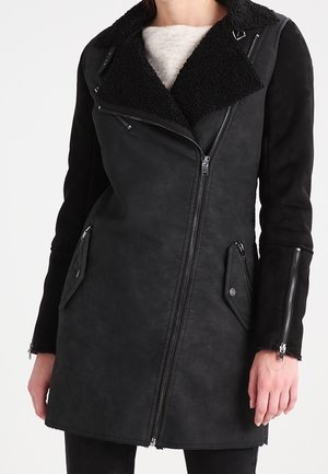 Manteau noir asymétrique à fermeture éclair avec col texturé, poignets zippés et poches à rabat inclinées, porté par une personne avec un pull léger en dessous.