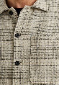 Chemise en tissu texturé beige avec des motifs en damier noirs, dotée de boutons noirs et d'une poche à rabat sur le côté gauche.