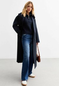 Marinblå lång kappa över en mörk tröja, parat med vida blå jeans och vita sneakers. En liten brun väska hänger från armen.
