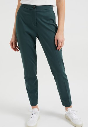 Femme portant un pantalon slim vert foncé et des baskets blanches, debout devant un fond clair uni.