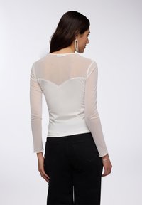 Maglietta trasparente a maniche lunghe di colore bianco, con un design aderente e un yoke posteriore scallopato. Indossata con pantaloni neri a vita alta.