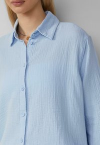 Helleblaues Button-Down-Hemd aus strukturiertem Stoff. Verfügt über einen Kragen, eine Knopfleiste vorne und einen lockeren Schnitt mit sichtbaren Nähten.