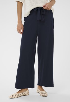 Femme portant un pantalon large marine à taille nouée, associé à des ballerines tissées crème et un pull en maille crème.