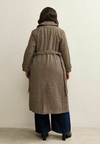 Długi płaszcz w kratę w brązowo-kremowych kolorach z paskiem, kołnierzowym dekoltem i bocznymi kieszeniami. Nosi się go z szerokimi dżinsami.