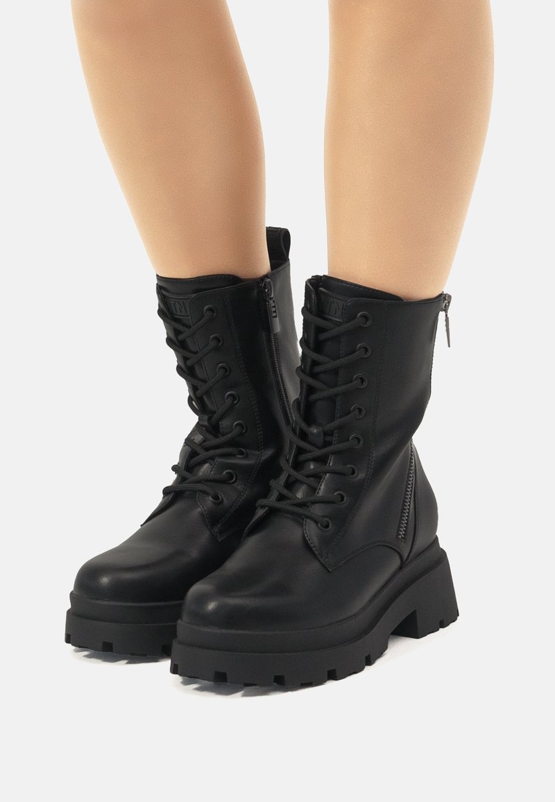 Botas de tobillo de cuero negro con suela gruesa, parte delantera con cordones y cremallera lateral. Textura suave con detalles de hardware en contraste.