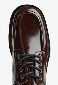 Scarpa in pelle lucida marrone scuro con suola nera spessa e testurizzata e lacci neri, mostrata dall'alto in primo piano su sfondo bianco.