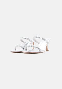 Ασημένια σανδάλια με τακούνι, πλεγμένα λουριά, διαφανείς λεπτομέρειες και μικρό τετράγωνο τακούνι. Λείο υλικό με λαμπερό φινίρισμα.