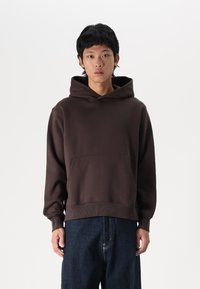 Brown hooded sweatshirt made from textured fabric, featuring a large front pocket and ribbed hem. Worn with dark denim jeans.