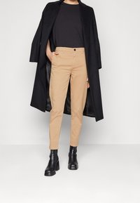 Beiga chinos med smal passform, svart långkappa och ankelstövlar. Kappan har en texturerad yta och en öppen design fram, vilket skapar en lager-på-lager look.