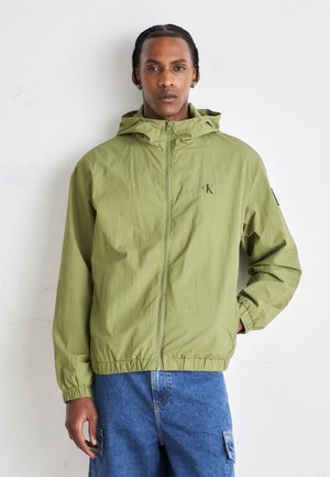 Giacca leggera verde chiaro con zip e cappuccio, polsini elasticizzati e un piccolo logo sul petto. Tessuto leggero con una finitura leggermente testurizzata.