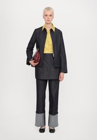 Giacca di jeans scuro con una camicia gialla a colletto, gonna abbinata e pantaloni larghi in jeans scuro con polsini grigi. Tiene una piccola borsa color maroon.