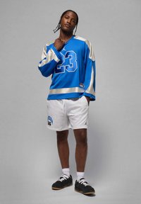 Jersey azul de manga larga con detalles plateados y el número 23. Combinado con pantalones cortos blancos que muestran el gráfico "Jordan 84". Zapatillas negras completan el conjunto.