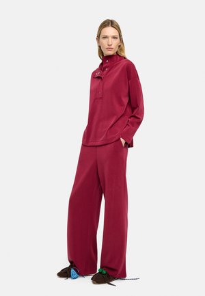 Kobieta stoi, ubrana w czerwony sweter z długim rękawem i zapiętym kołnierzykiem na guziki, dopasowane szerokie spodnie oraz ciemnobrązowe mokasyny w stylu owiniętym.