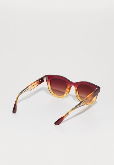Lunettes de soleil avec une monture bicolore, supérieure rouge et inférieure ambre, dotées de verres ronds et d'une finition lisse et brillante. Les branches vont de l'ambre au rouge.