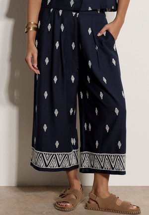 Marineblaue, weit geschnittene, gekürzte Hose mit weißen geometrischen Mustern und Rand, getragen mit braunen Sandalen mit offenen Zehen und goldenen Armreifen am linken Handgelenk.