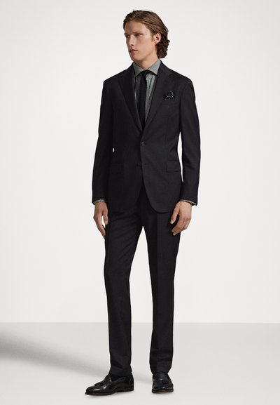 Czarny, dopasowany garnitur o smukłym kroju, z klapami wciętymi, wzorzystym krawatem i poszetką. Całość dopełniają błyszczące czarne eleganckie buty.