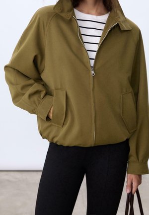 Frau trägt eine olivgrüne Jacke mit Reißverschluss über einem weiß-schwarz gestreiften Shirt, kombiniert mit schwarzen Hosen, hält einen braunen Taschenhenkel.