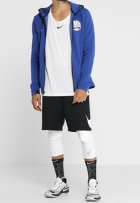 Sudadera con capucha azul con cremallera y logotipo, camiseta blanca, pantalones cortos negros, leggings blancos, calcetines negros con estampado y zapatillas deportivas grises.