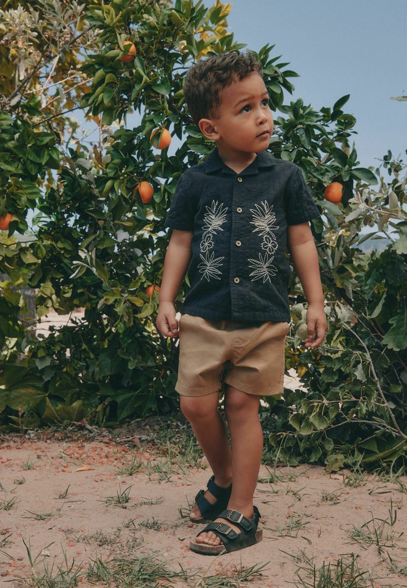 Jeune garçon en chemise noire brodée et short beige debout sur un sol sablonneux proche d'un arbre à oranges portant des fruits mûrs.