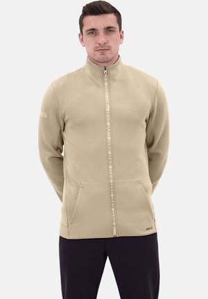 Beige ritsjack van zachte stof, met een hoge kraag, twee voorvakjes en een gemerkte rits met herhaalde tekst.
