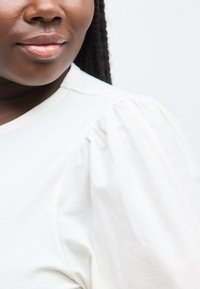 Κοντινό πλάνο στο κάτω μέρος του προσώπου και τον ώμο μιας γυναίκας που φοράει λευκό τοπ με φουσκωτά μανίκια, σε απλό ανοιχτό φόντο.