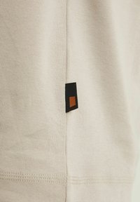 Tan T-shirt featuring a ribbed texture, detailed stitching, and a black and orange fabric tag on the side.