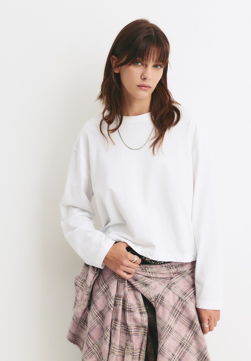 Weißes langärmliges T-Shirt aus Baumwolle, kombiniert mit einem rosa karierten Hemd, das um die Taille gebunden ist. Schlichte, lockere Passform mit minimalen Details.