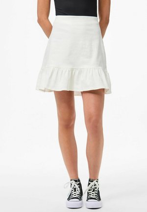 Jupe blanche évasée avec un ourlet froncé, fabriquée en tissu léger. Associée à des baskets montantes noires avec des semelles et des lacets blancs.