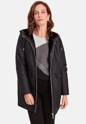 Zwarte gewatteerde parka met een ritssluiting aan de voorkant, twee voorzakken en een interieur met nepbont. Daaronder een grijs en zwart gebreid vest met strepen.
