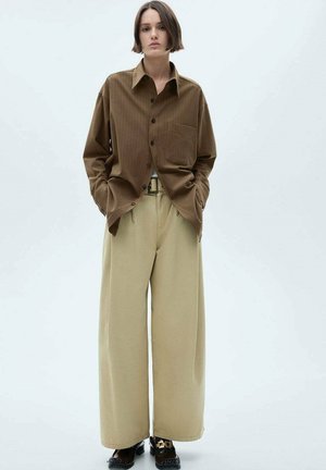 Camicia a righe verticali marroni con un taschino, abbinata a pantaloni beige larghi e calzature nere. Design semplice e comodo.