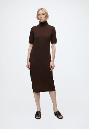 Femme aux cheveux blonds courts portant une robe midi marron à col roulé et des sandales à talons ouvertes noires, debout devant un fond blanc uni.