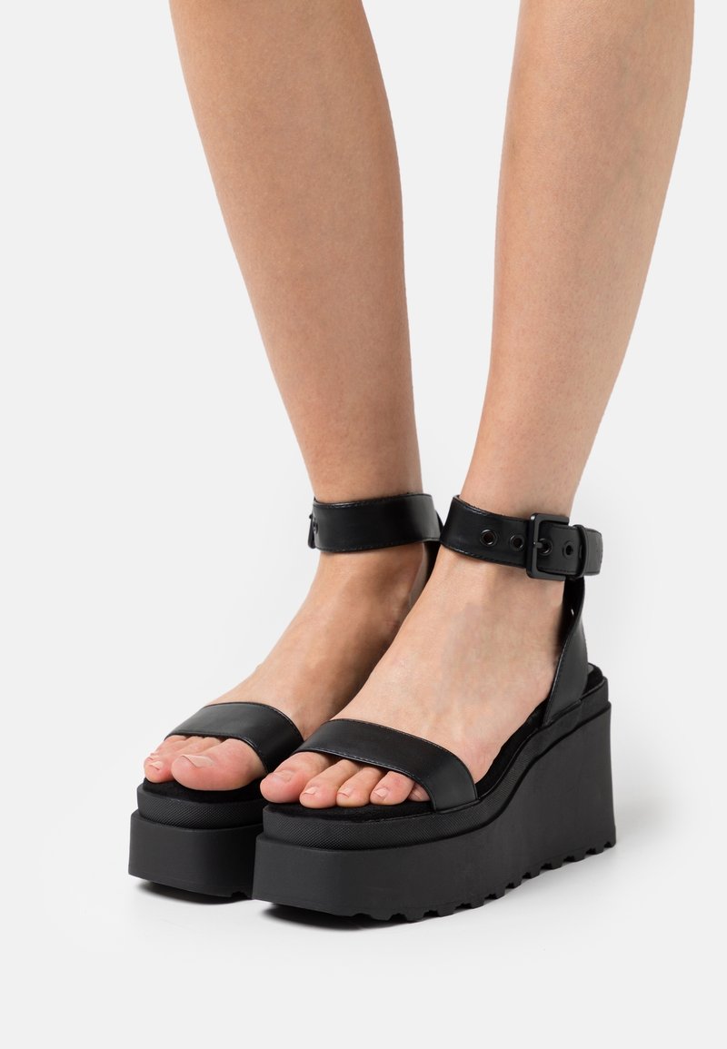 Personne portant des sandales compensées noires avec brides à la cheville et semelles épaisses, debout sur un fond blanc. Seules les jambes inférieures et les pieds sont visibles.