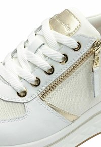 Sneaker bianco con tessuto testurizzato, accenti metallici dorati, cerniera in metallo dorato e lacci bianchi. Presenta una punta arrotondata e un profilo basso.