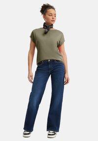 Oliwkowa bluzka z krótkim rękawem i szerokim dekoltem, zestawiona z ciemnoniebieskimi spodniami szerokimi w nogawkach. Czarny szalik jest związany wokół szyi.