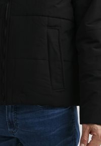 Sort pufferjakke med en quiltet tekstur, der har en sidelomme og en lynlås. Bæres over blå denimjeans.
